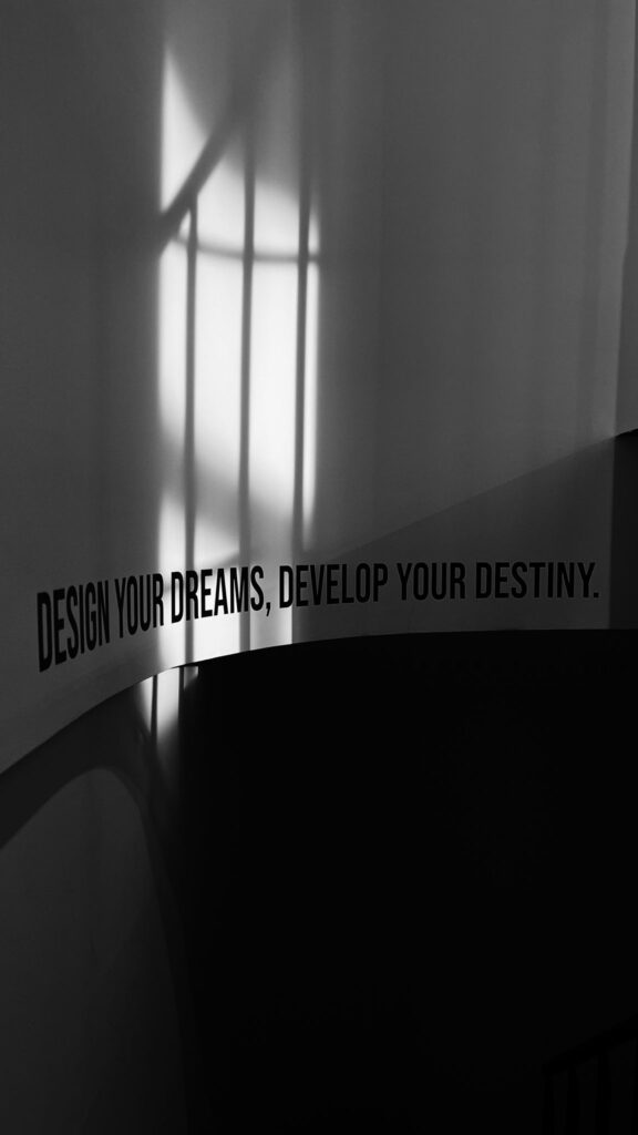 Een artistieke zwart-witfoto die de schaduw van een raamkozijn op een gebogen wand laat zien, met de inspirerende tekst 'DESIGN YOUR DREAMS, DEVELOP YOUR DESTINY.' in zwarte letters onderaan de afbeelding. De lichtval en schaduw creëren een rustige, contemplatieve sfeer.